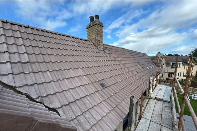 A finished roof in new concrete interlocking tile, running up to a stone chimney, photographed from the scaffold.