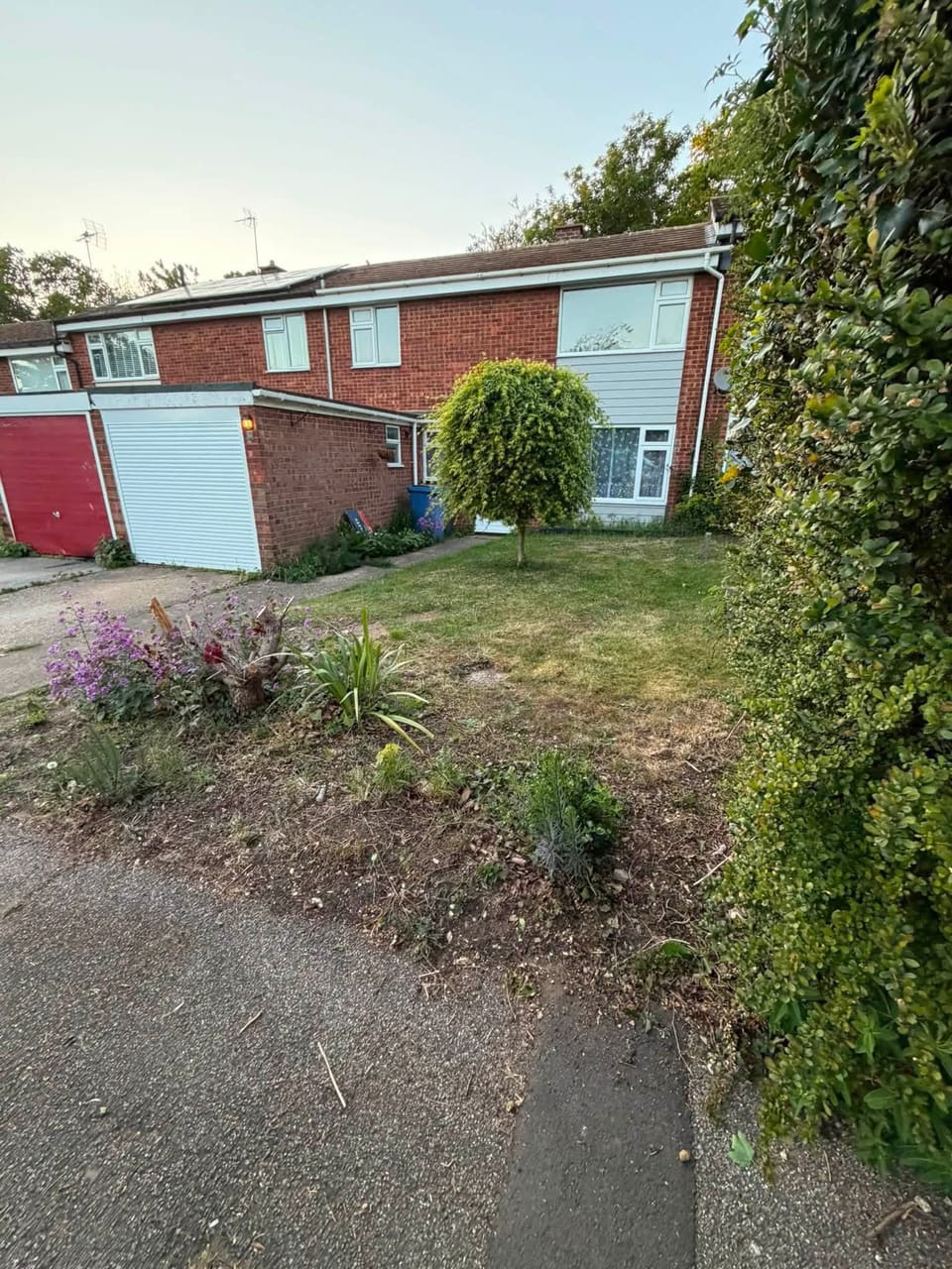 The frontage from the side before the work: unmade ground and a patchy verge beside a red garage door.