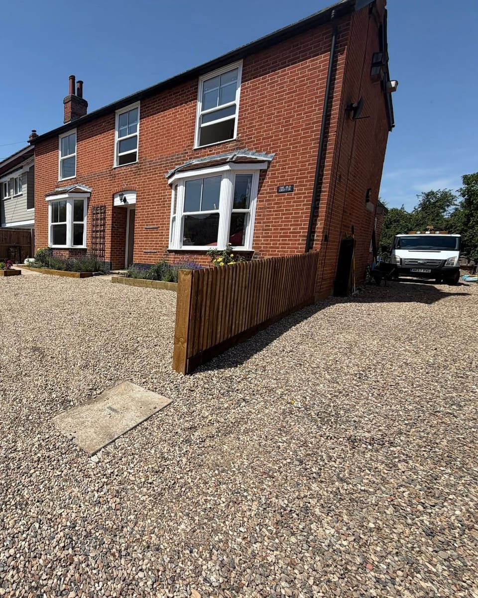 A new close board timber fence beside a gravel driveway at the front of a red brick house.