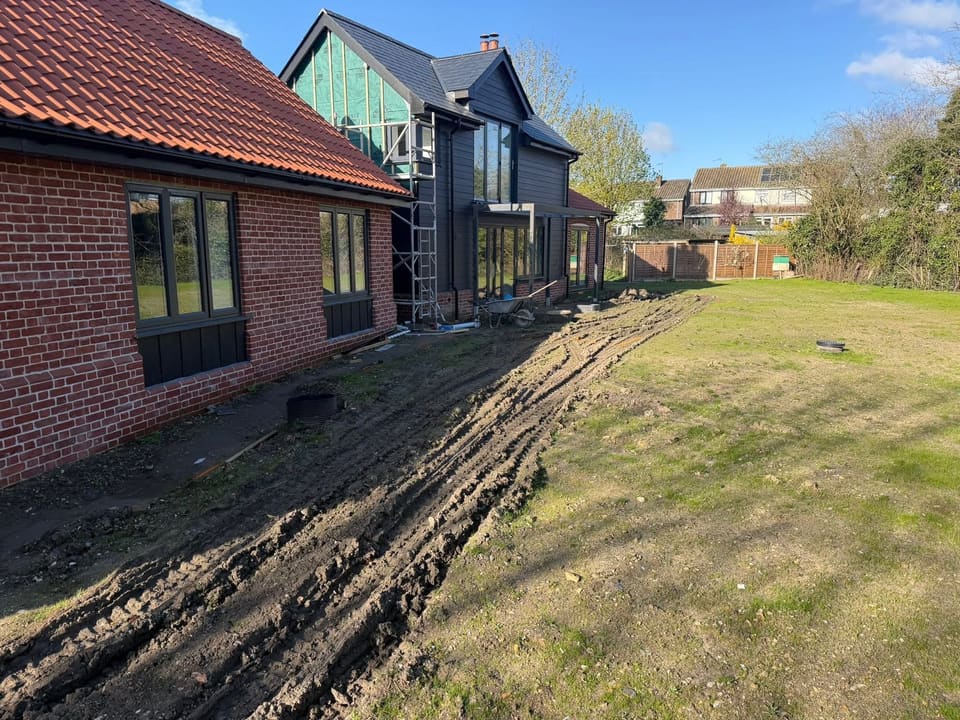 Churned ground and tyre ruts running the length of a house, with a wheelbarrow and an open manhole.