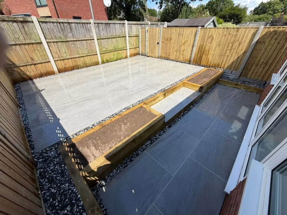 A patio halfway through: the first porcelain slabs down, the sleeper edge going in, the gravel margin waiting.