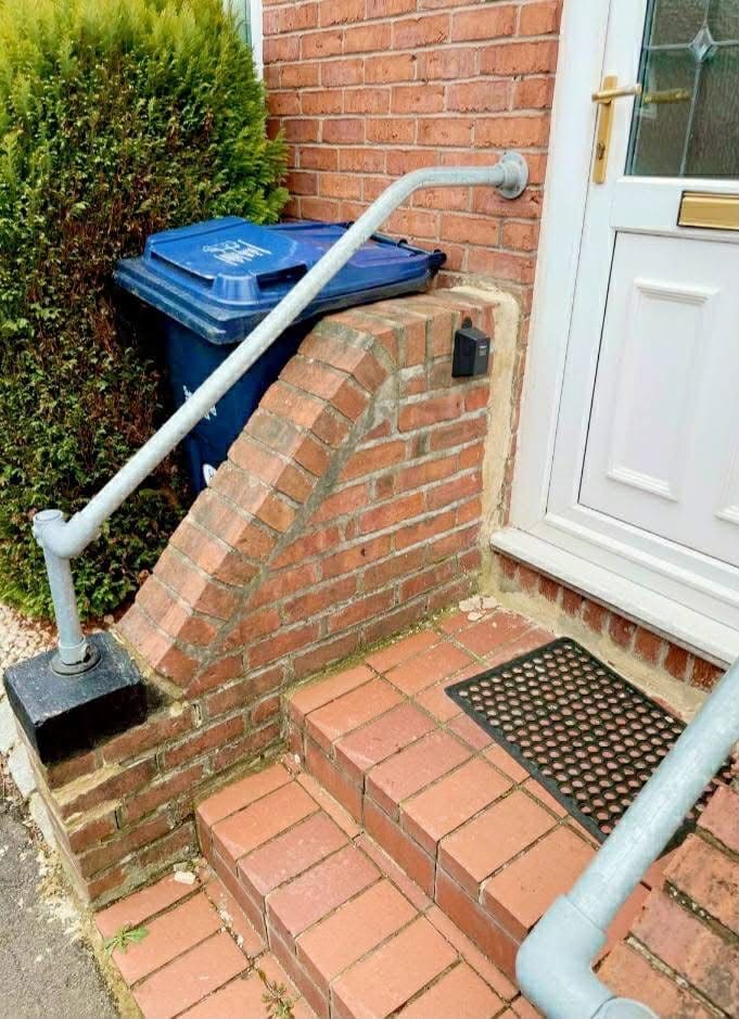 Brick steps and a raked cheek wall at a front door, with a galvanised handrail and a blue recycling bin beside it.