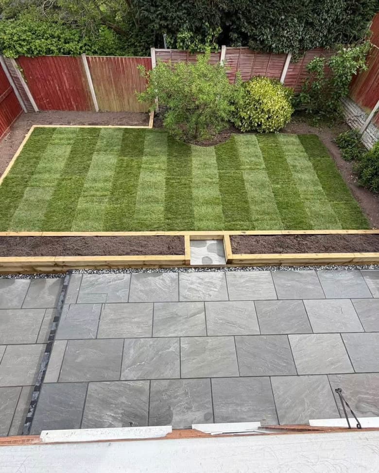 A finished back garden: grey riven porcelain paving with a gravel margin, a timber sleeper edge, and freshly laid striped turf behind it.
