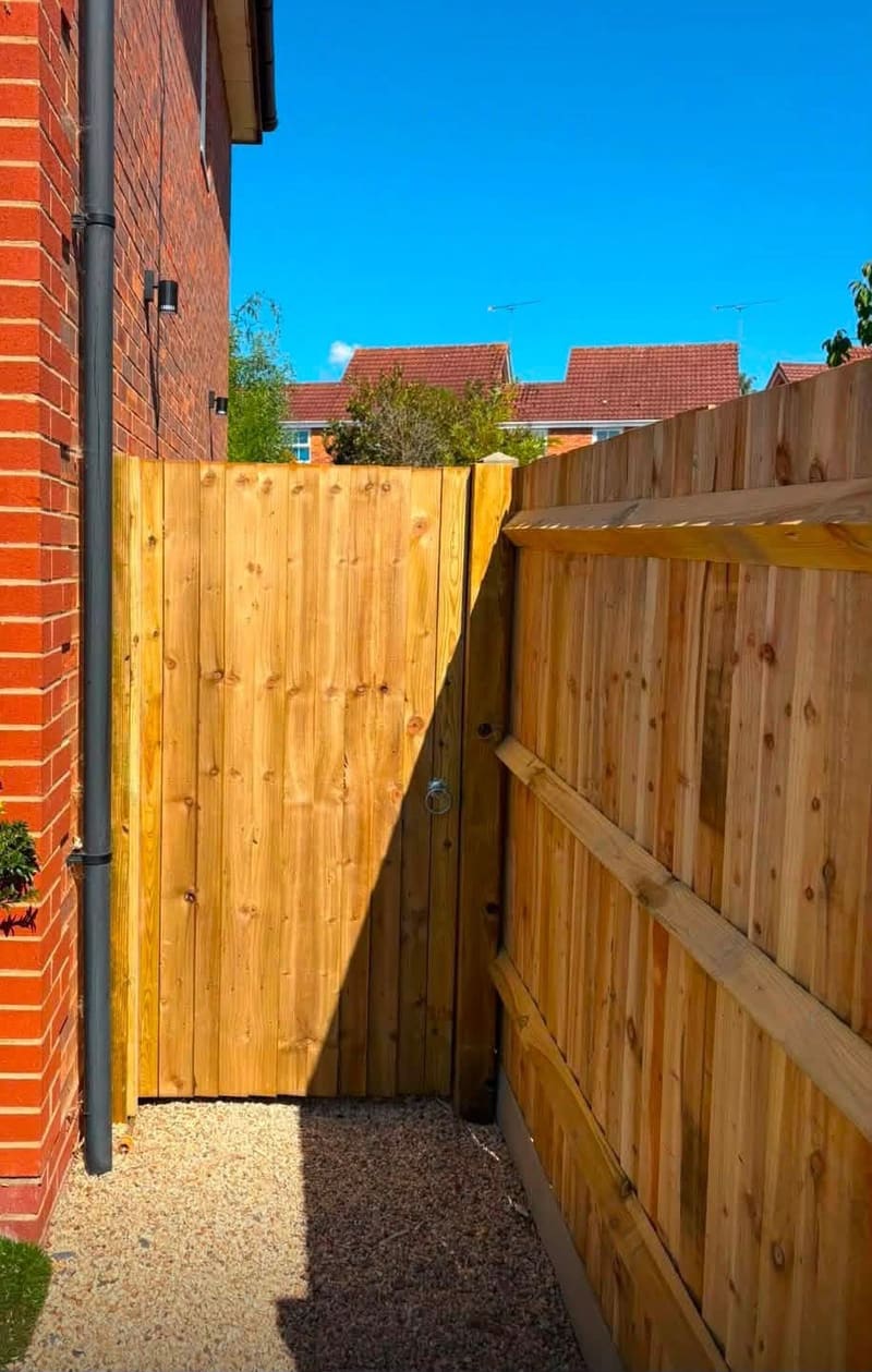 A new close board timber fence and a matching gate between a red brick house and the boundary, over a gravel path.