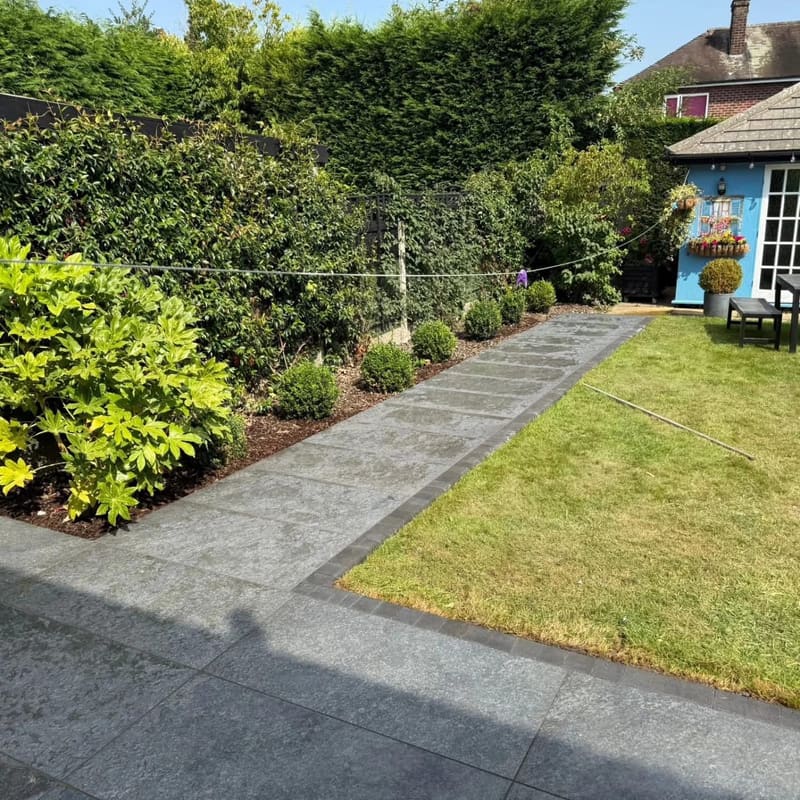 A wide grey plank paving path laid across a lawn to a blue summerhouse, with a planted border of clipped shrubs alongside it.