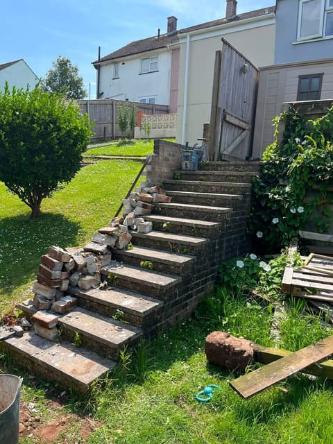 Old concrete garden steps up a slope, with loose bricks stacked along the edge and grass grown over the treads.