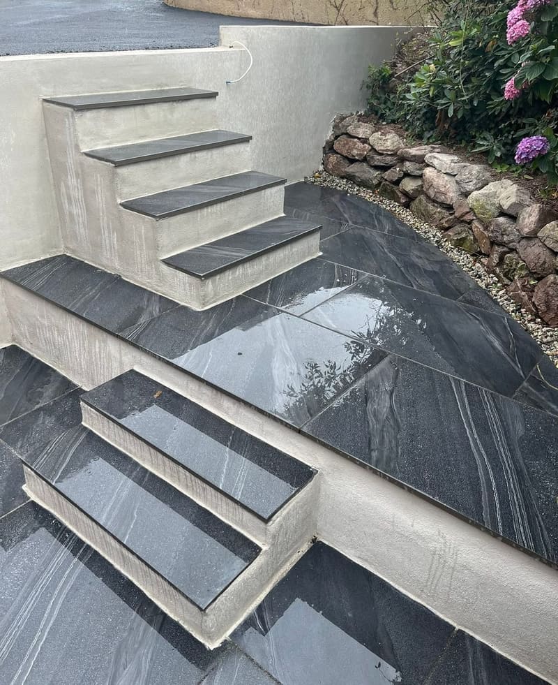 Steps and a landing in dark porcelain with rendered cheeks, beside a stone retaining wall and a hydrangea in flower.