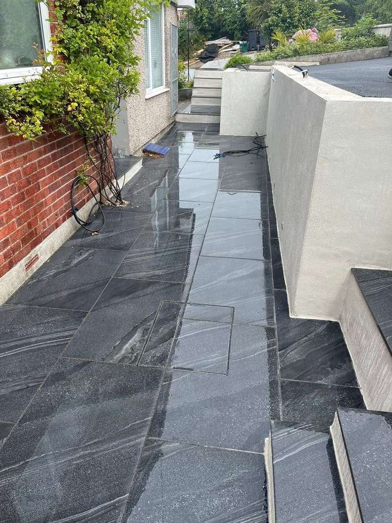 A finished dark porcelain terrace running alongside a house, wet from rain, with a new white rendered retaining wall on one side.