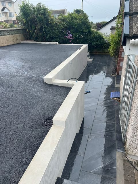 New black tarmac meeting a dark porcelain terrace, with a white rendered retaining wall running between them and a railing beyond.