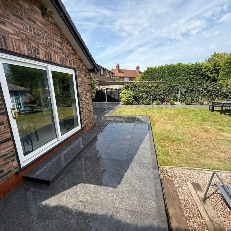 A dark porcelain patio outside white sliding doors on a red brick house, wet, running out to the lawn with a gravel margin at the edge.