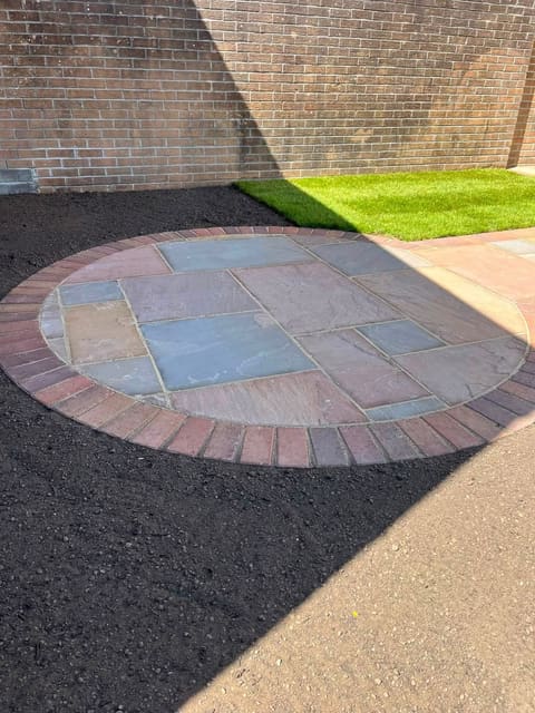 A circular sandstone feature with a red brick ring around it, set against a brick wall, with dark soil and grass beside it.