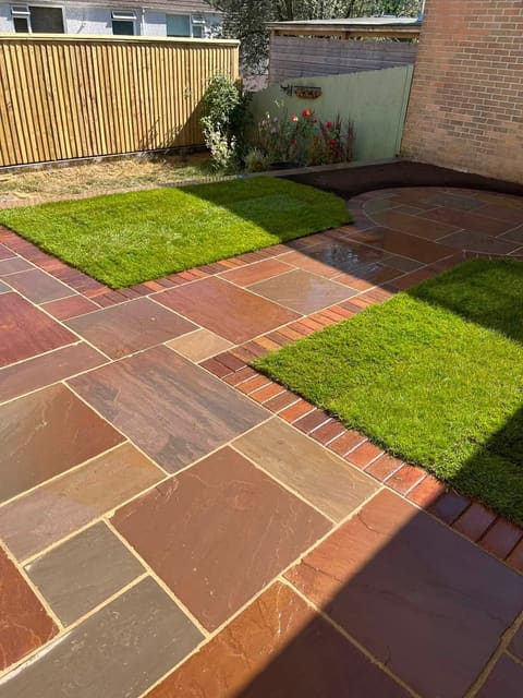 Riven sandstone paving with red brick edging crossing between two squares of turf, against a timber fence and a brick wall.