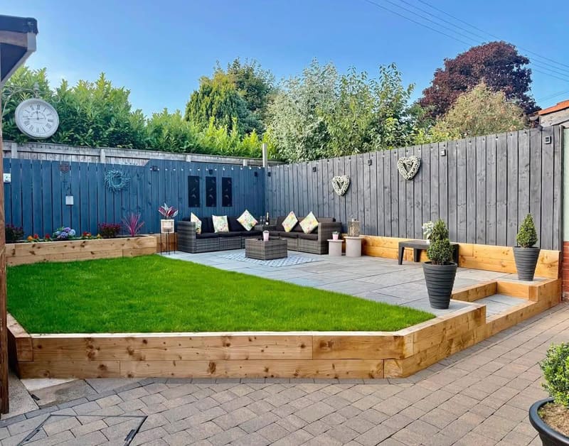 A finished garden from the corner: grey paving, a timber sleeper raised edge holding a lawn, block paving and rattan furniture against a dark grey fence.