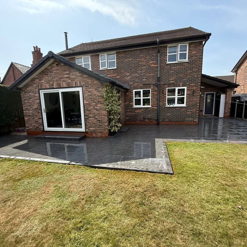 The back of a two storey brick house with a new dark porcelain patio, wet from rain, edged and stepped down to the lawn.