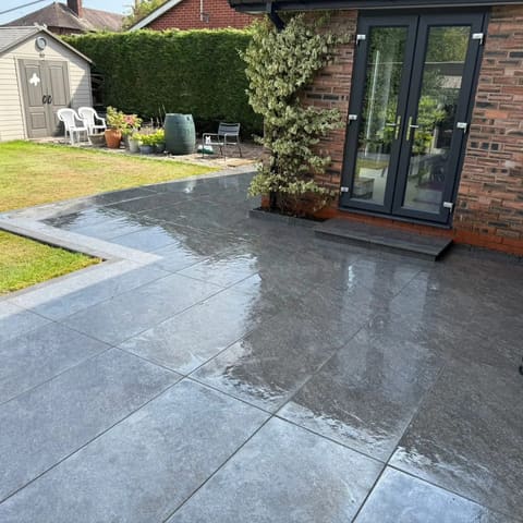 Large dark porcelain slabs, wet, running up to grey french doors with a step, a shed and a hedge beyond.