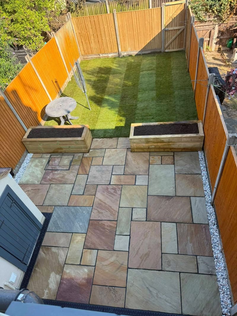 A finished back garden: riven sandstone paving in mixed sizes, two timber sleeper planters and new striped turf, inside an orange close board fence.