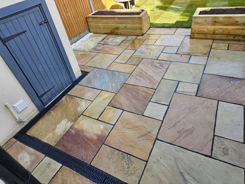 Riven sandstone paving in mixed sizes running up to a dark timber gate, with a black slot drain along the edge and a sleeper planter behind.