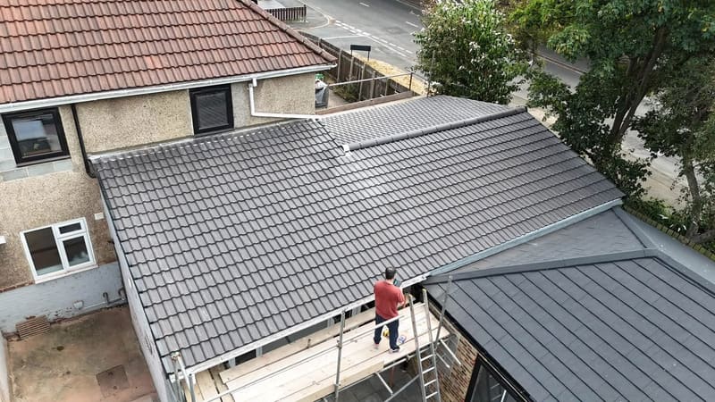 A flat pitched roof re-covered in new grey interlocking tile, seen from above, with a roofer standing on the scaffold board at the eaves.