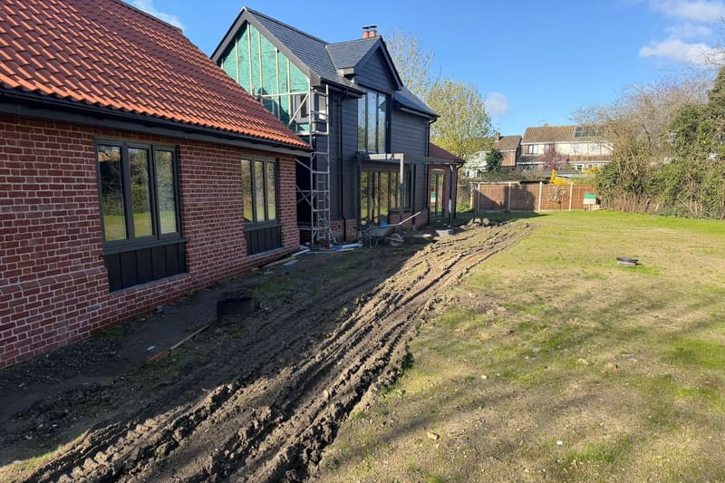 Churned ground and tyre ruts running the length of a house, with a wheelbarrow and an open manhole.