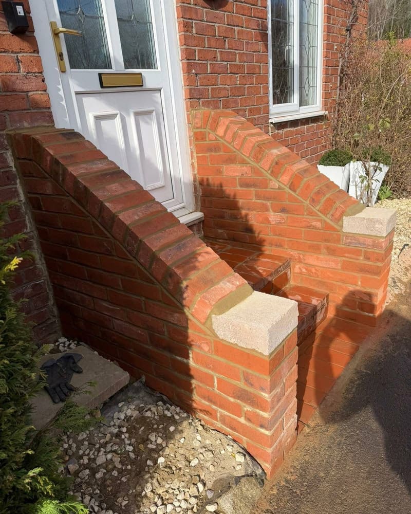 A raked brick cheek wall being built beside a front door, with the ground beneath it open and gravel showing.