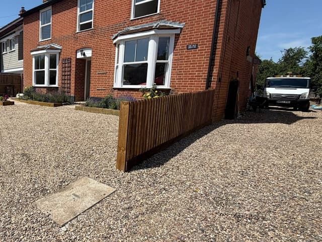 A new close board timber fence beside a gravel driveway at the front of a red brick house.